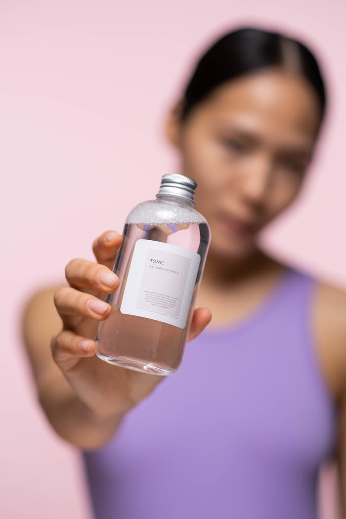 A woman presents a skincare tonic in a studio setting with blurred background.